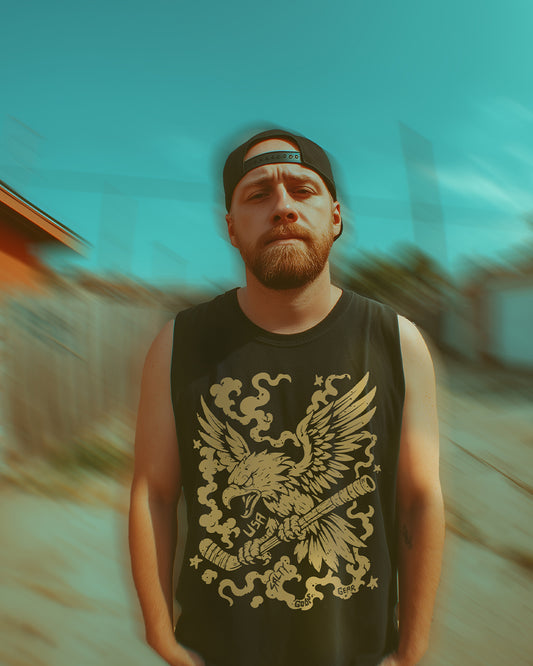 Man wearing a black muscle tank top with a bold gold eagle and hockey stick graphic. The man has a red beard, is wearing a black snapback hat backwards, and is standing outdoors in an urban setting with a teal sky and blurred background.