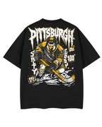 A black short-sleeve T-shirt shown flat against a white background, featuring a large back graphic of a hockey player in a Pittsburgh design with the word 'Pittsburgh' in a jagged white font and additional graphic text and accents in white and yellow.