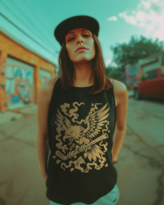 Woman wearing a black muscle tank top with a bold gold eagle and hockey stick graphic. She has straight brown hair, a black flat-brim cap worn forward, and is standing in an urban alleyway with graffiti and a red truck in the background. The sky is teal with scattered clouds and the image has a dramatic, street-style vibe.