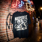 Black t-shirt with 'Nashville' design on a brick wall with neon signs in the background