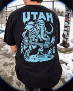 Person wearing a black t-shirt with blue 'UTAH' and graphic design outdoors.