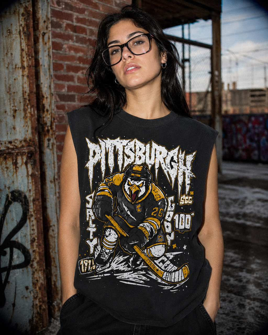 A woman wearing a black sleeveless graphic t-shirt with a Pittsburgh hockey design, standing in front of a rustic brick and metal wall.