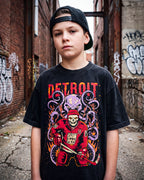 A young man wearing a black Detroit  graphic t-shirt and a black baseball cap, standing in an urban alleyway with brick walls and graffiti.